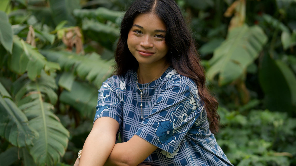 Woman in a blue patterned dress squatting in a grassy area with green foliage.